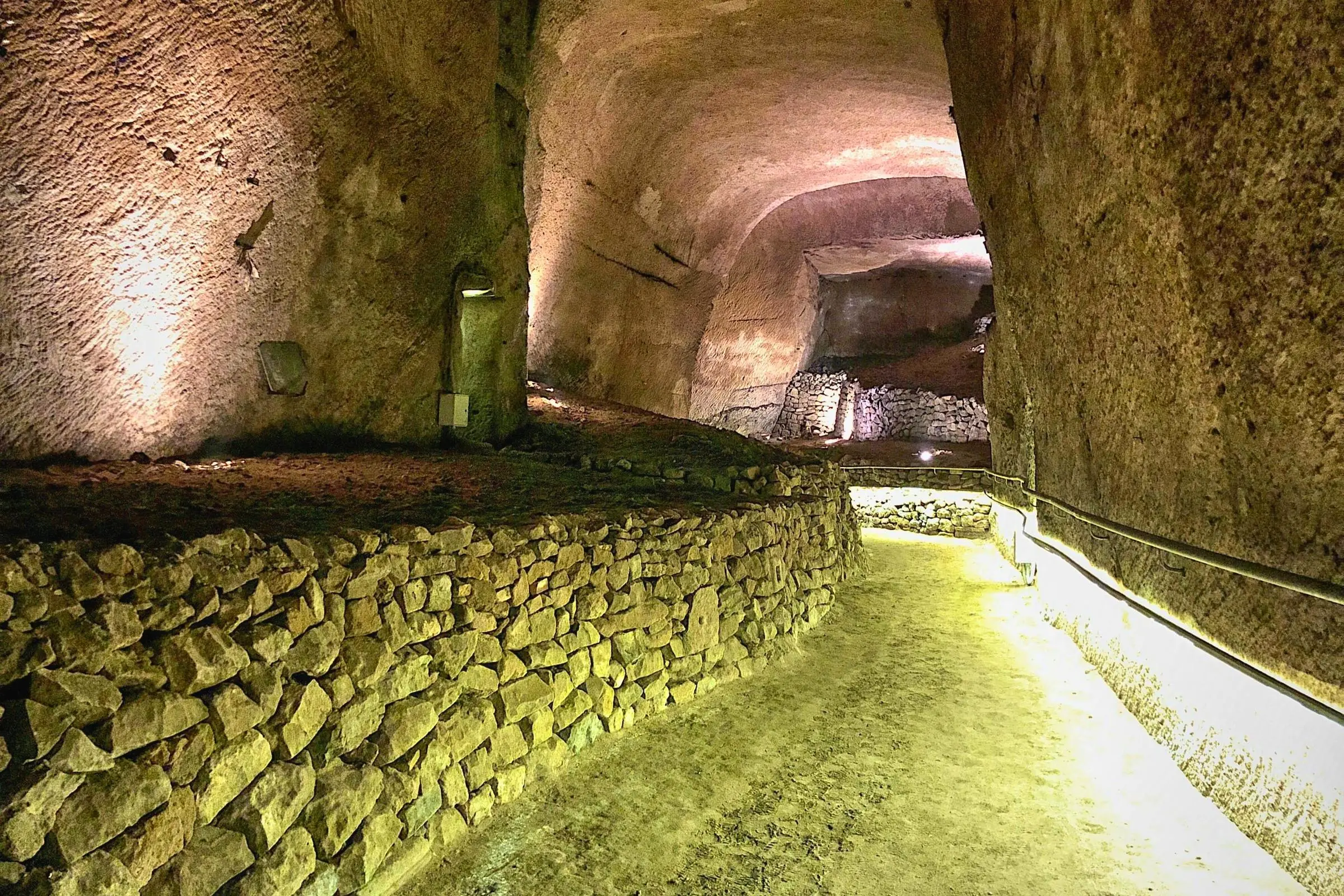 A wide, illuminated stone pathway winding through a high-vaulted tuff rock cavern with a low stacked-stone wall